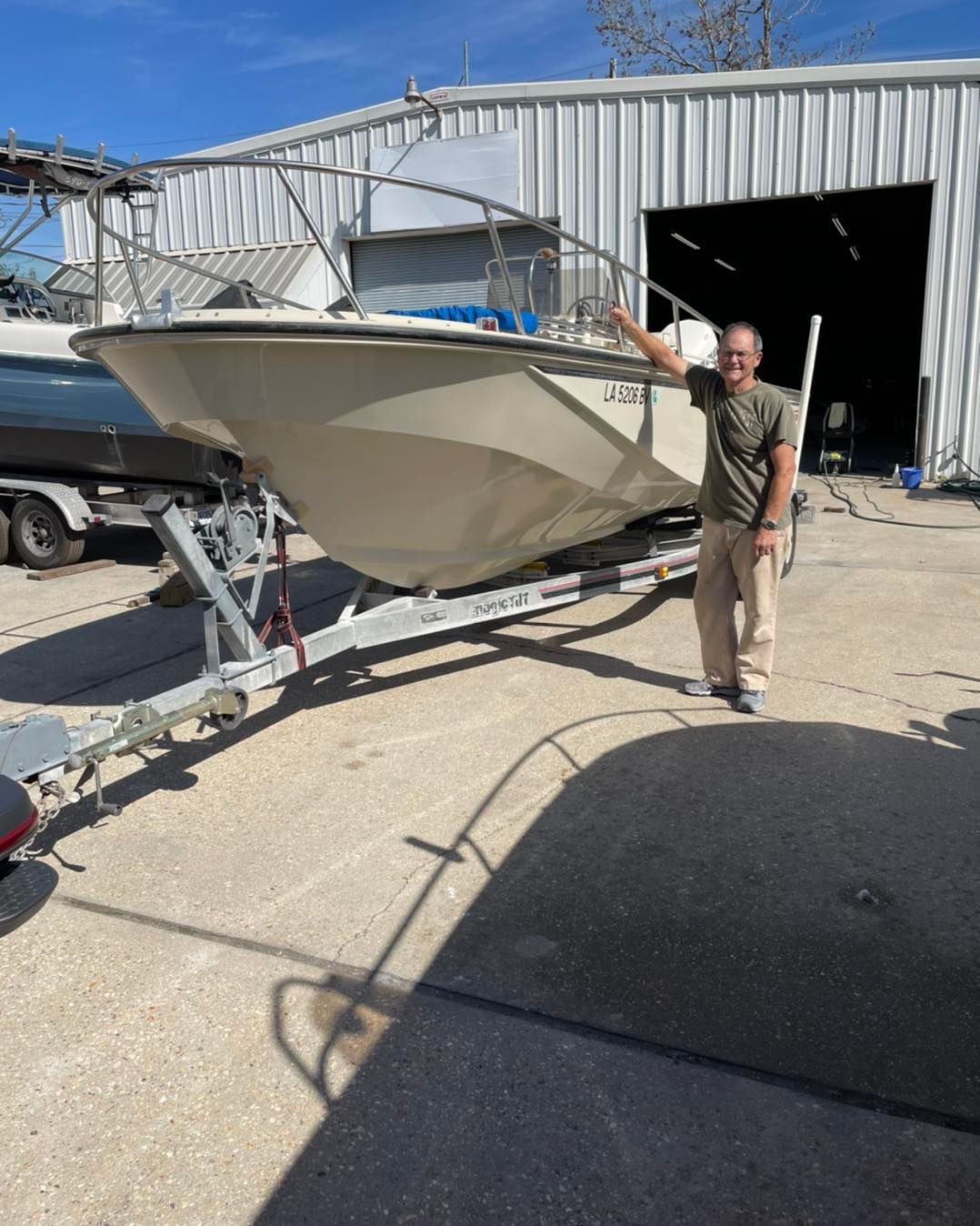 Man standing beside a beige boat on a trailer outside a metal workshop.