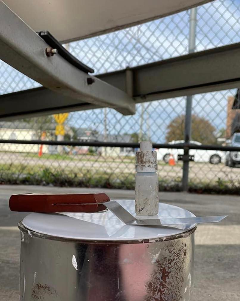 Metal pot on a table with a wooden-handled lid, under a covered outdoor area with a chain-link fence behind it