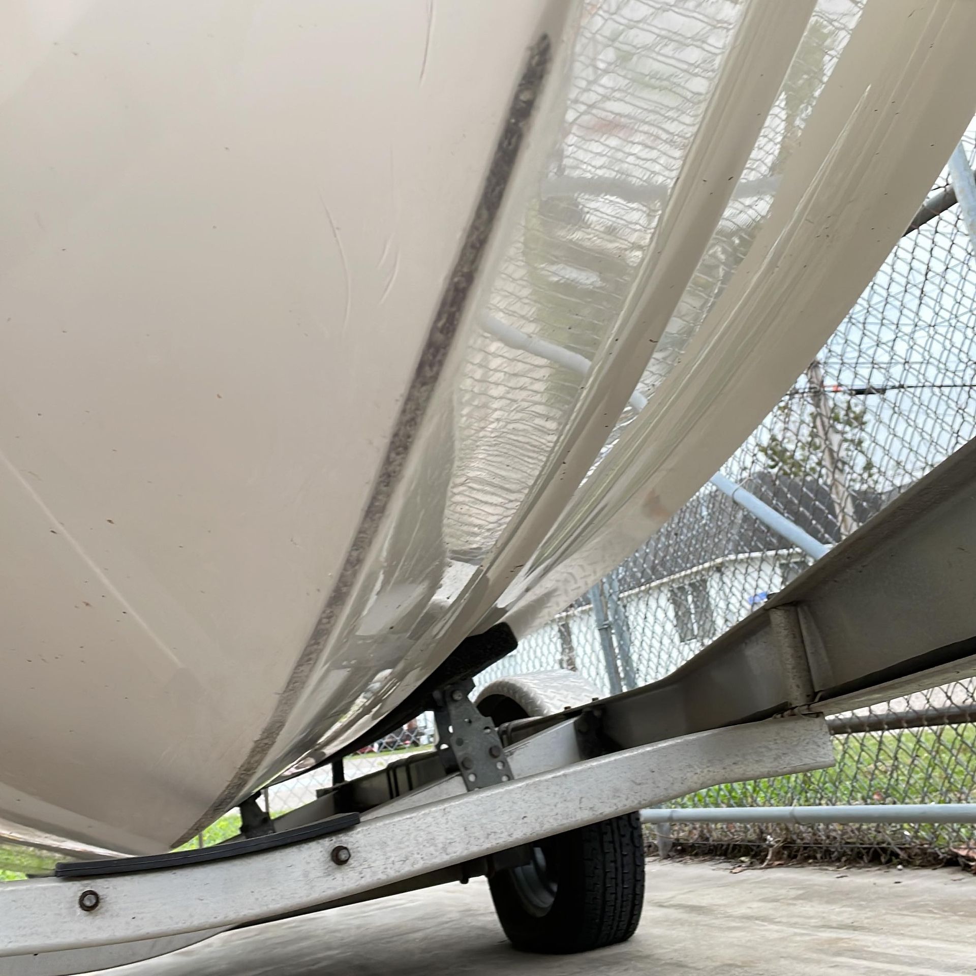 Boat on a trailer viewed from below, showing the hull and metal frame beside a chain-link fence.