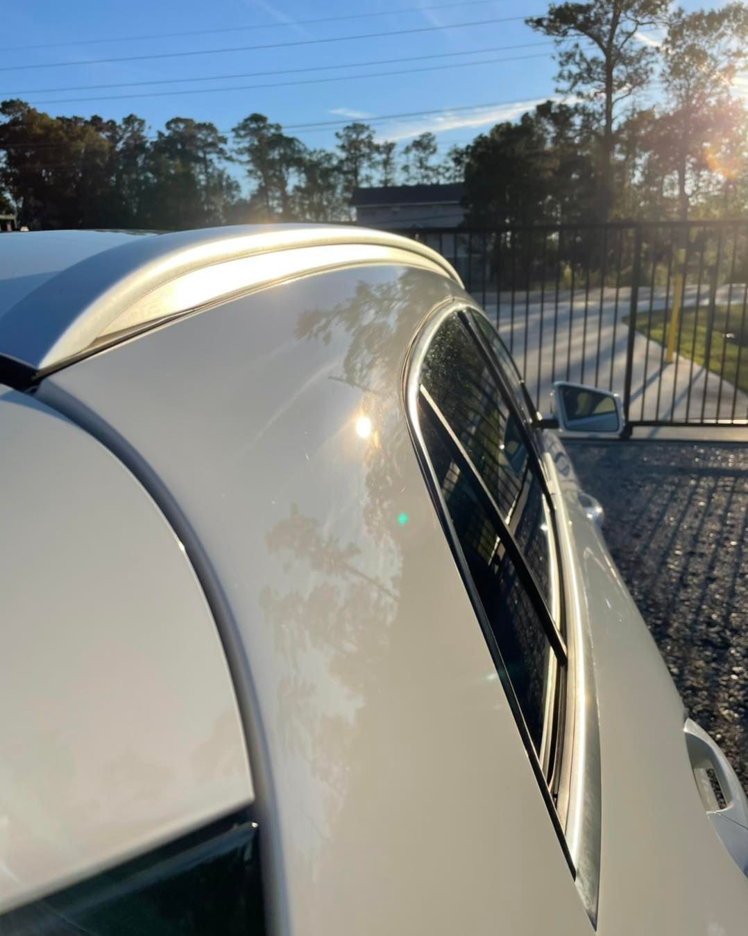 White car roof and rear window in sunlight beside a gated driveway and trees