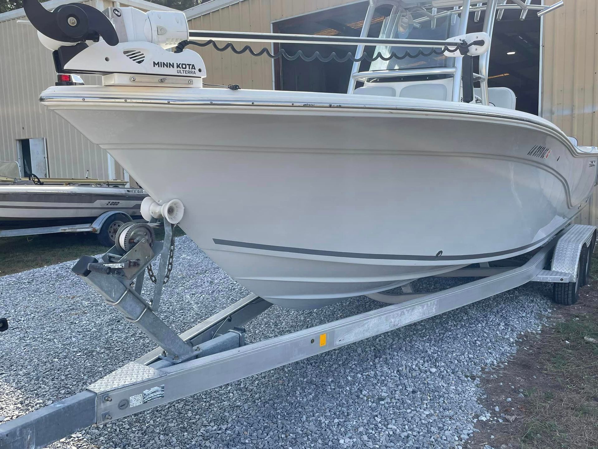 White boat on a trailer parked on gravel beside a building, with a seat and railing visible.