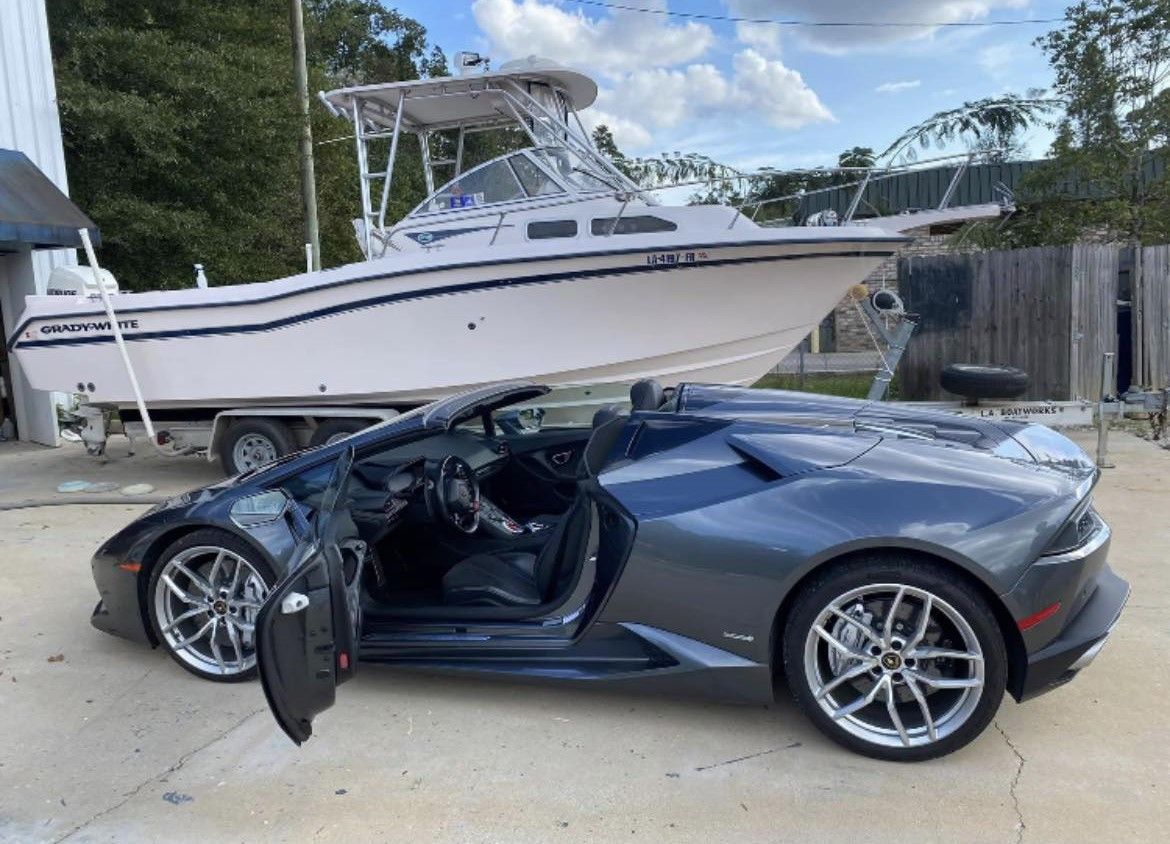 Dark gray sports car with driver door open parked beside a white boat on a trailer