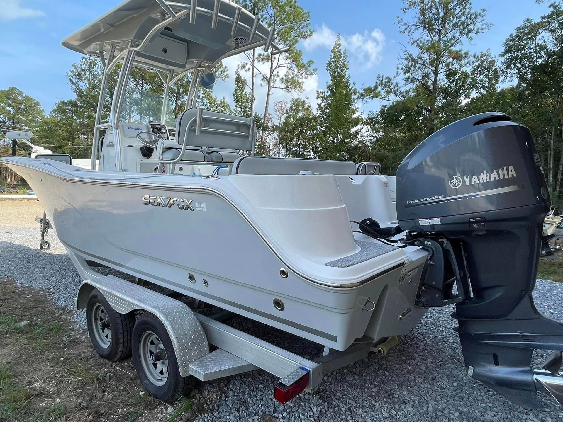 White center-console fishing boat on a trailer with twin outboard motors, parked outdoors under trees