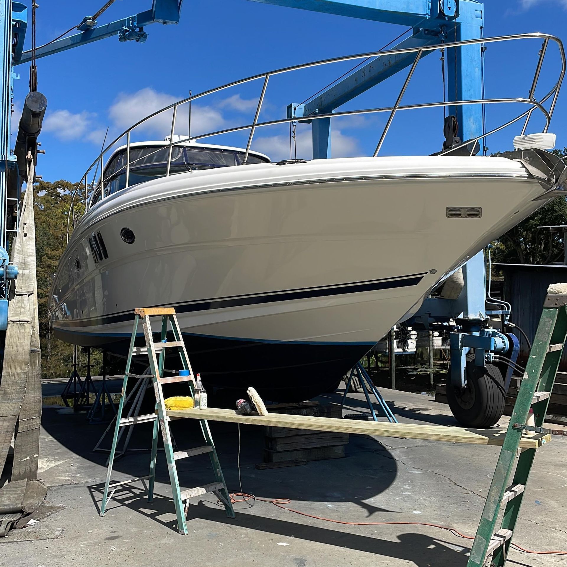 White sailboat on stands in a boatyard, surrounded by ladders and maintenance equipment under a blue sky