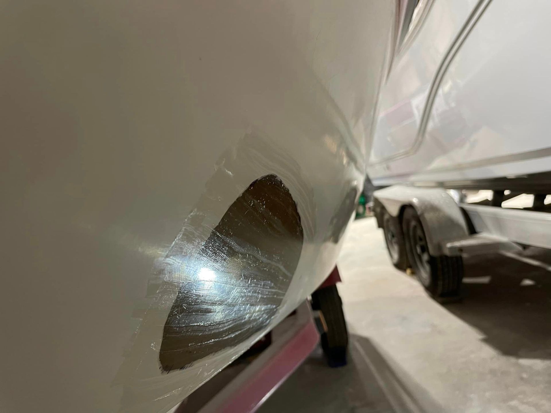 Close-up of a white boat hull with a damaged patch and another boat on a trailer in the background
