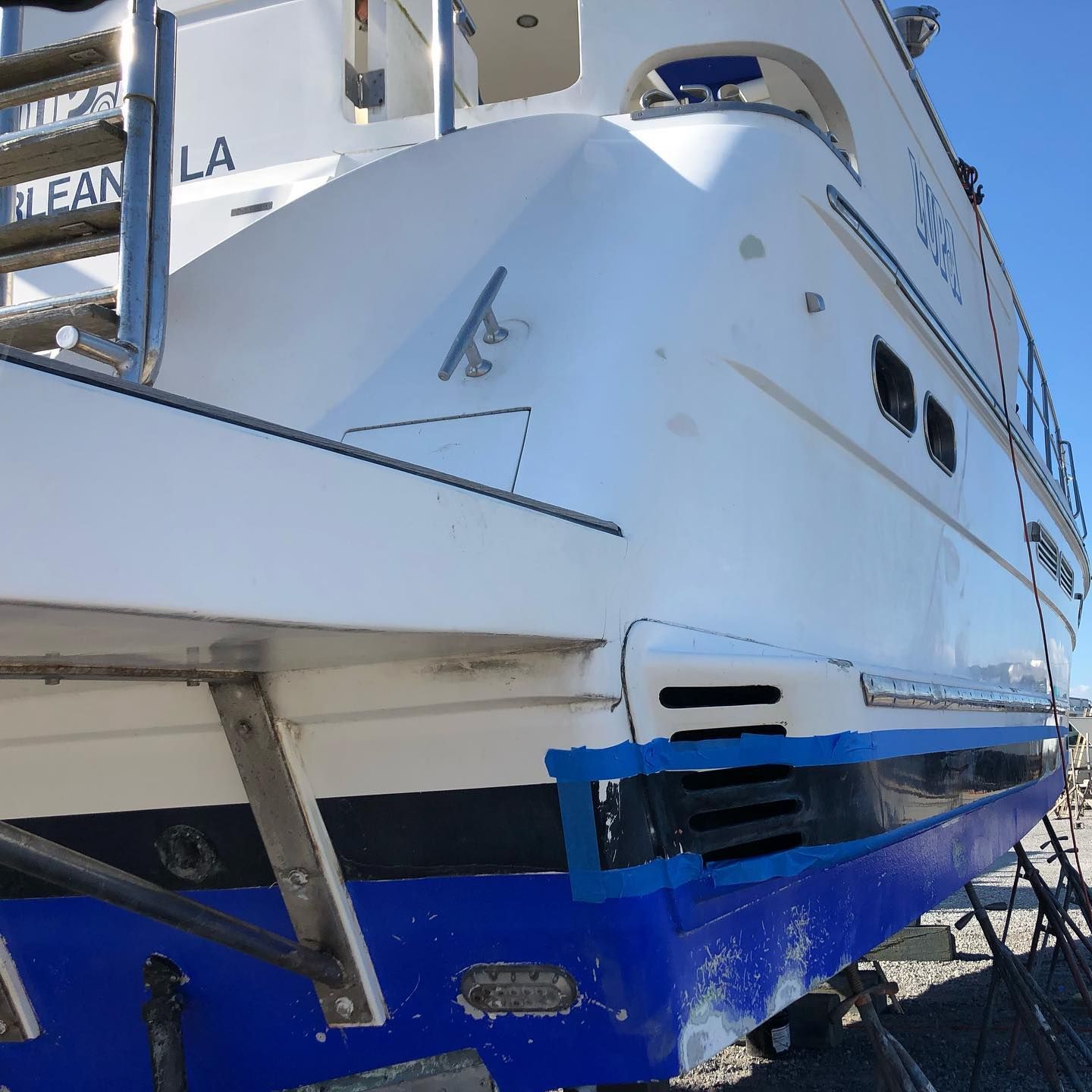 White and blue yacht hull at a dock, viewed from the side and front in bright sunlight