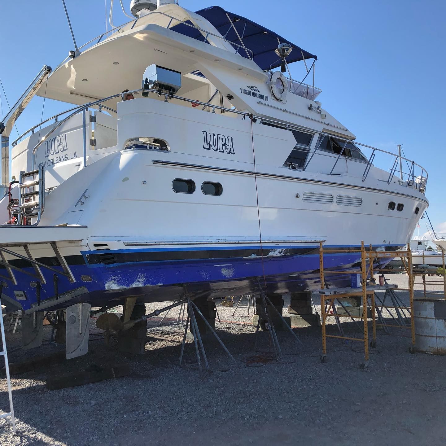 White yacht on supports in a dry dock, with a blue lower hull under a clear sky