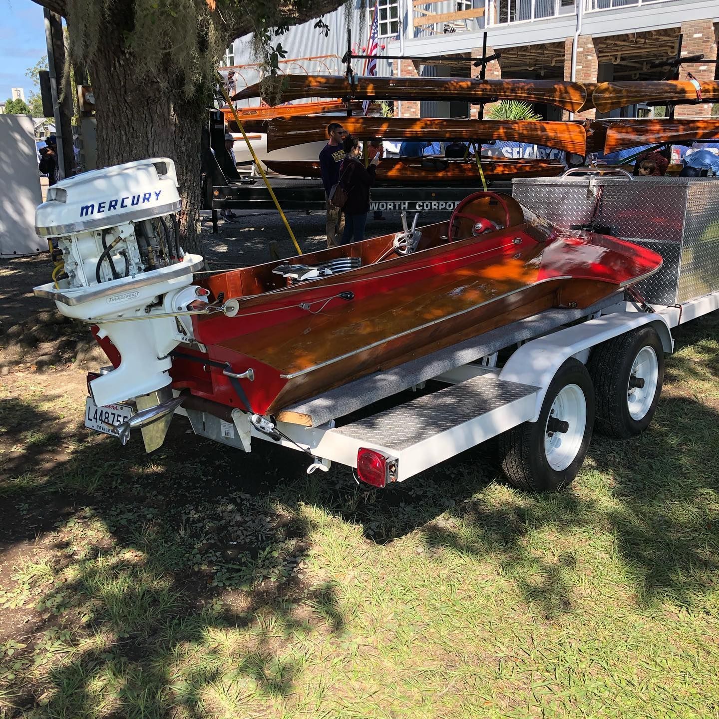 Red fishing boat on a trailer in a shaded grassy yard, with an outboard motor and orange kayaks stacked behind.