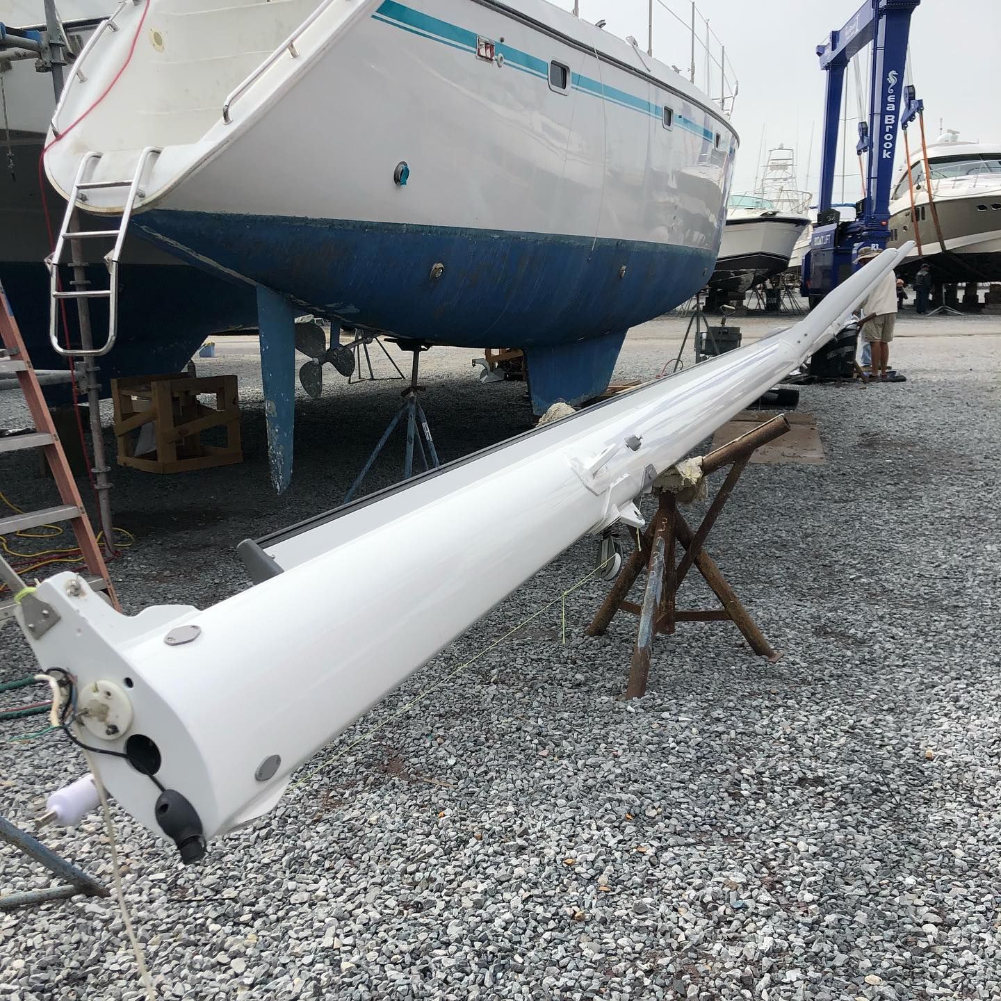 White sailboat mast lying on gravel beside a boat hull in a boatyard