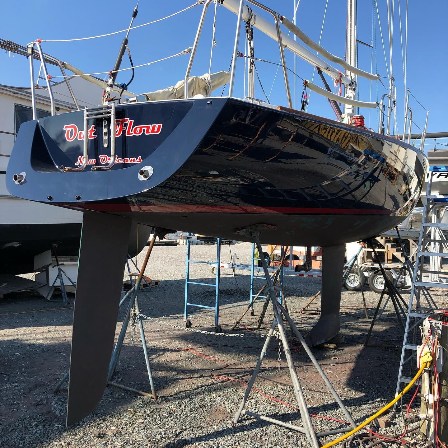 Black sailboat on stands in a boatyard, with a red-and-white name painted on the stern.