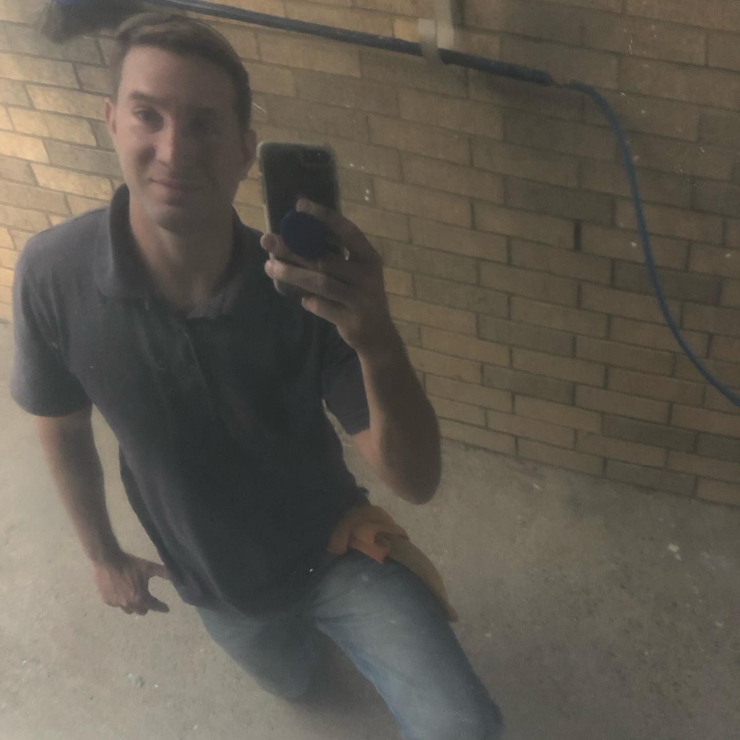 Person taking a mirror selfie in a brick-walled hallway, wearing a dark polo shirt and jeans.