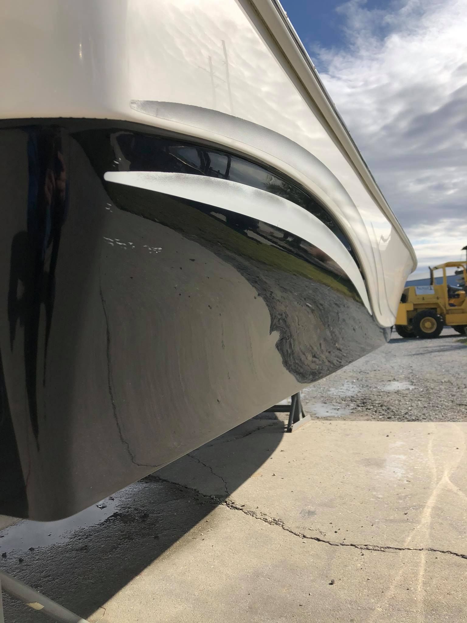 Close-up of a glossy black boat hull with white trim at a marina, with gravel and a truck in the background