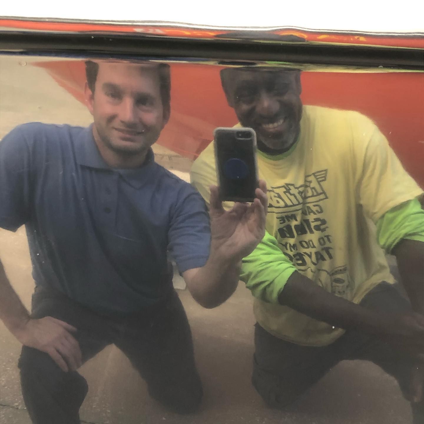 Two men crouch for a mirror selfie, one holding a phone, in front of a red vehicle.