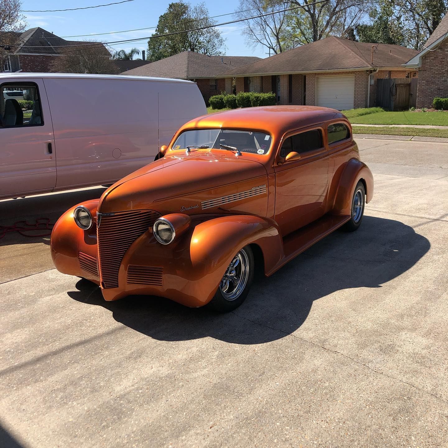 Orange vintage coupe parked on a residential street beside a white van on a sunny day