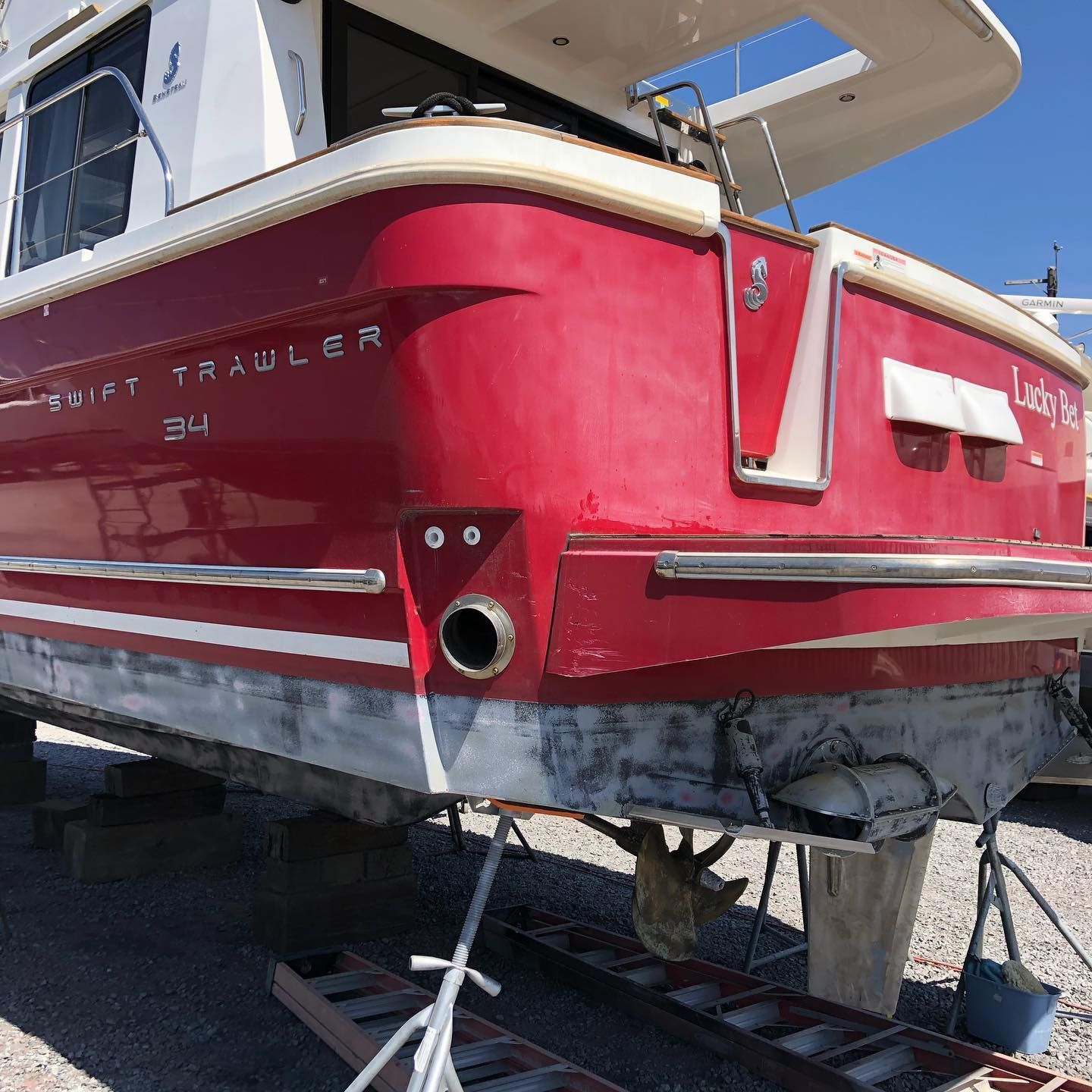Red-and-white sailboat on a trailer in a boatyard, viewed from the stern under a clear blue sky