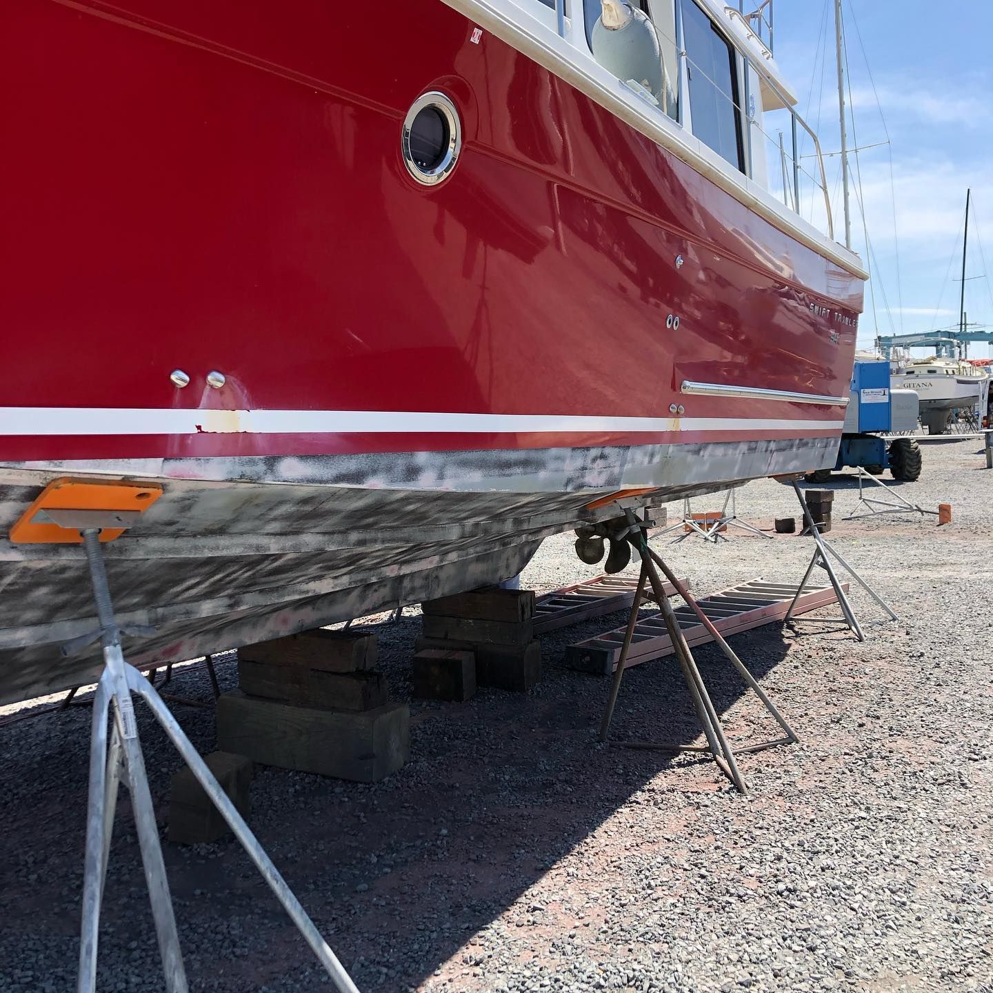 Red boat hull on stands in a gravel boatyard, with lower edge being repaired or painted