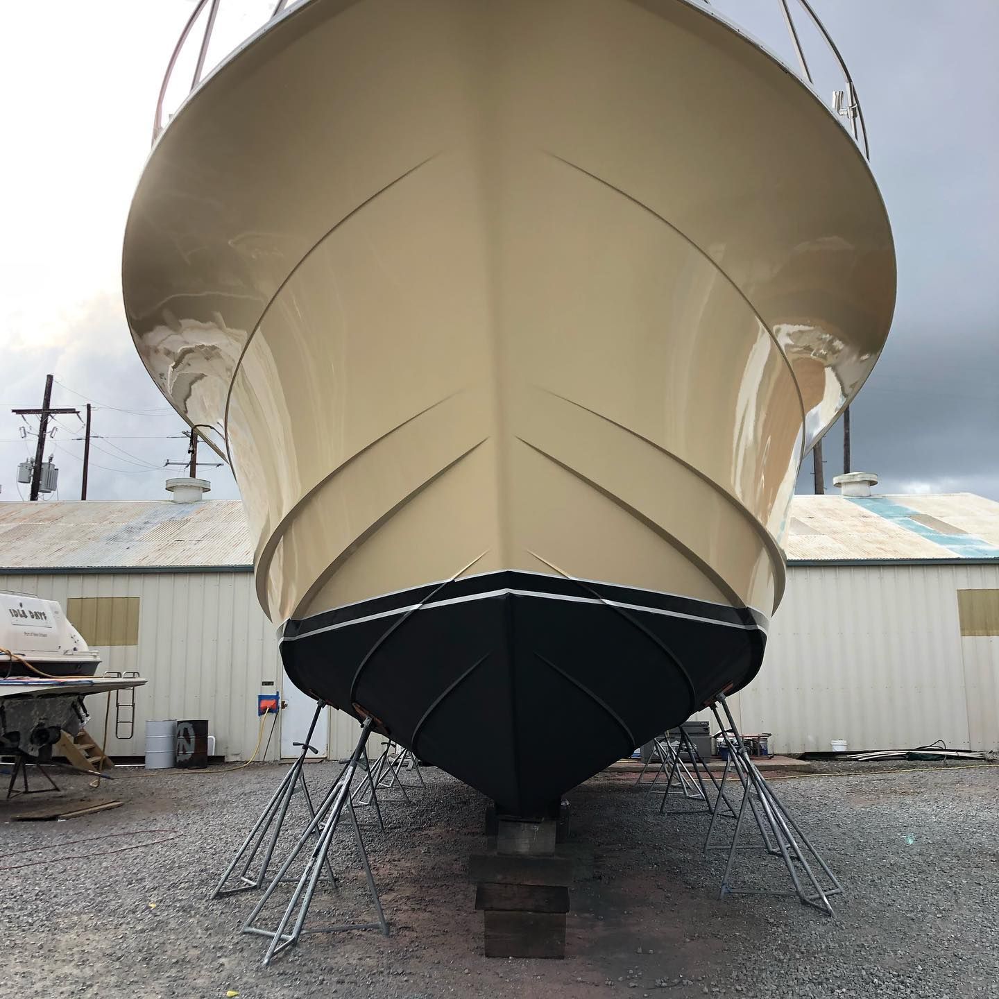Front view of a large beige sailboat on stands in a boatyard, with a black hull bottom.