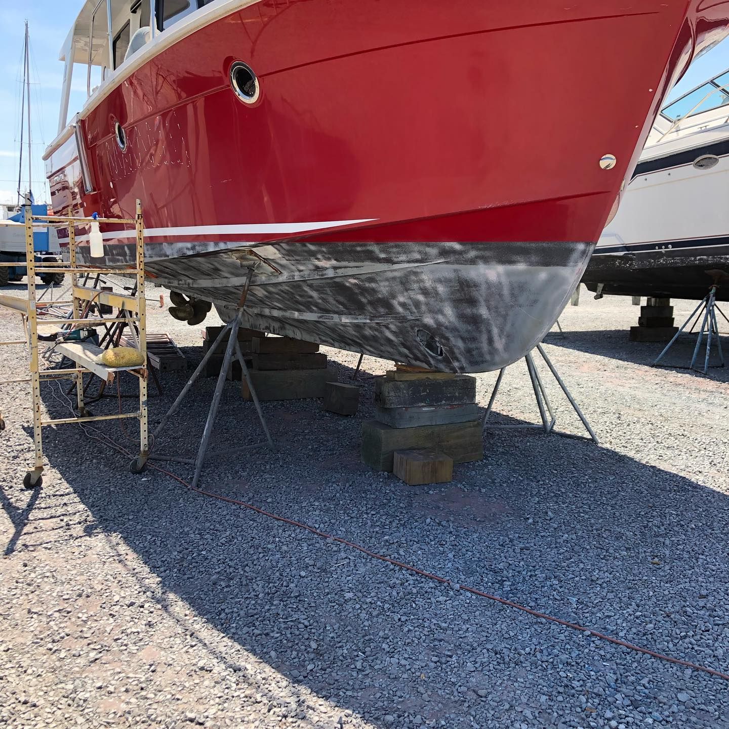 Red sailboat hull on supports in a gravel boatyard, with a black antifouling bottom.