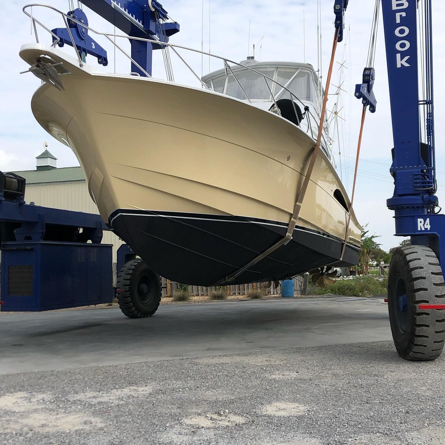 Large cream-colored boat being lifted by a blue crane in a boatyard, viewed from below