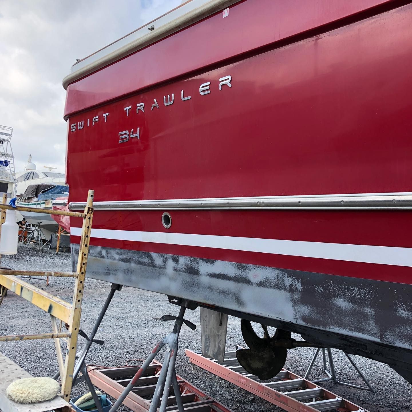 Red boat trailer on a gravel lot, with white trim and a yellow ladder beside it.
