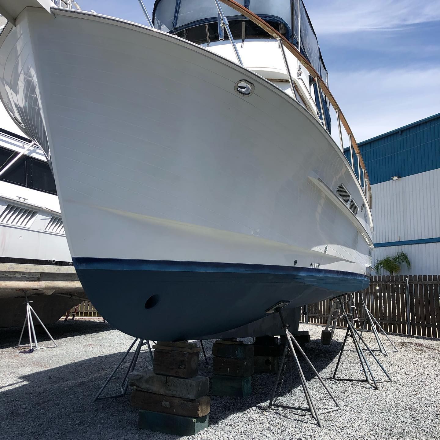 White sailboat hull on stands in a boatyard, with blue lower hull and nearby docked boats.