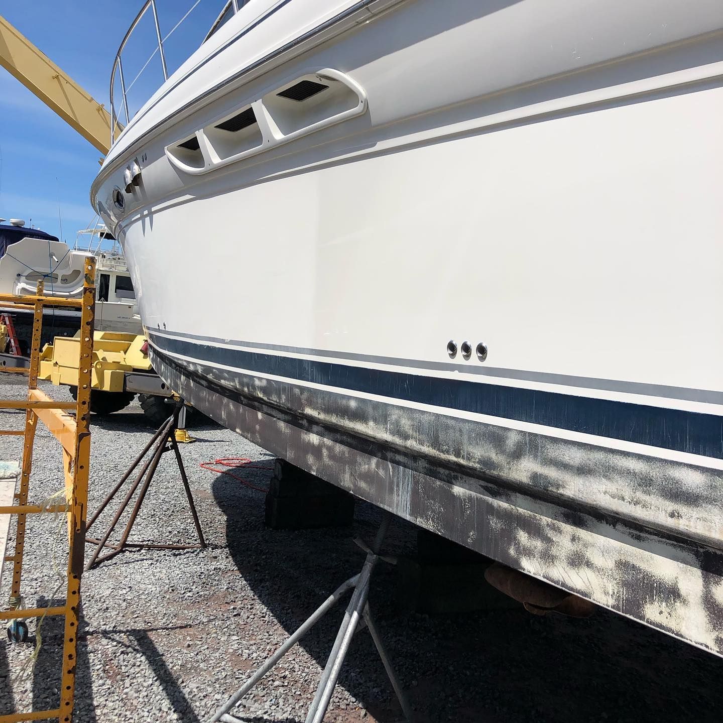 White boat hull on stands in a boatyard, with blue stripe and weathered lower side under bright sun