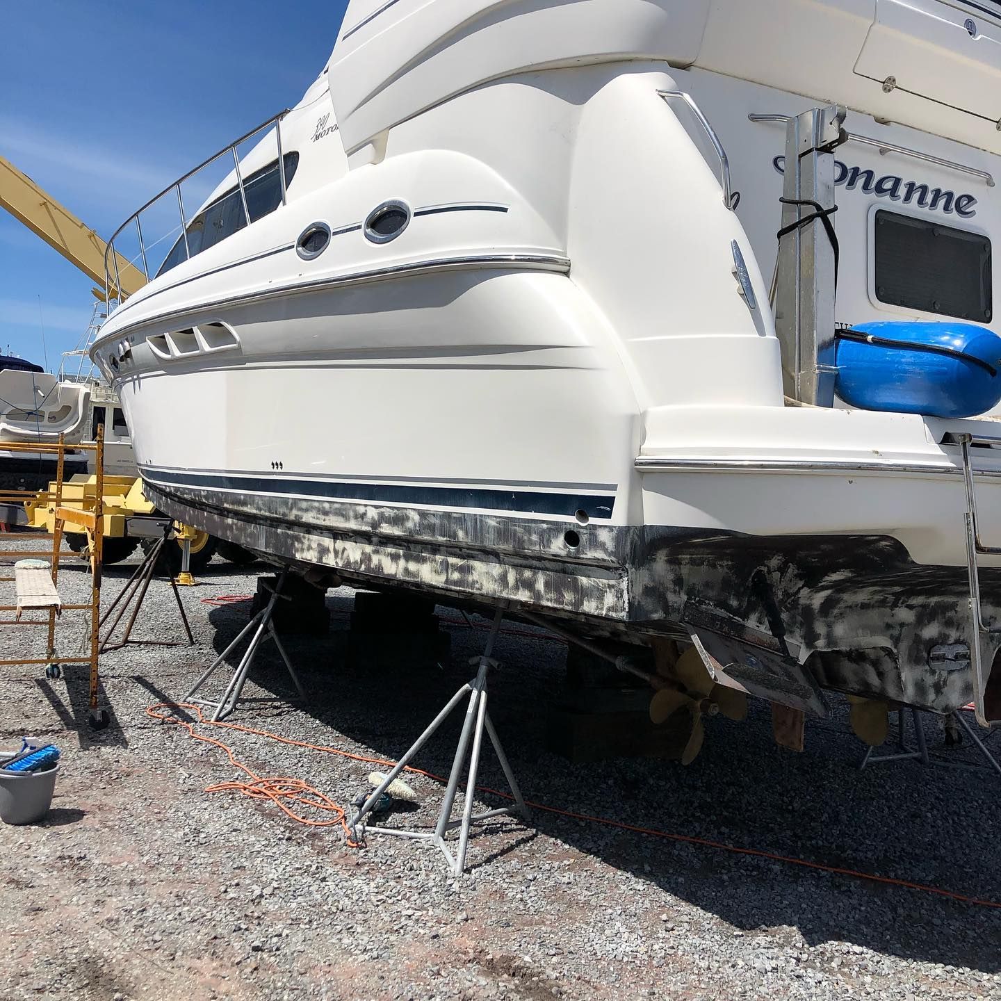 White yacht on stands in a boatyard, with a blue fender and visible hull damage or repair work.