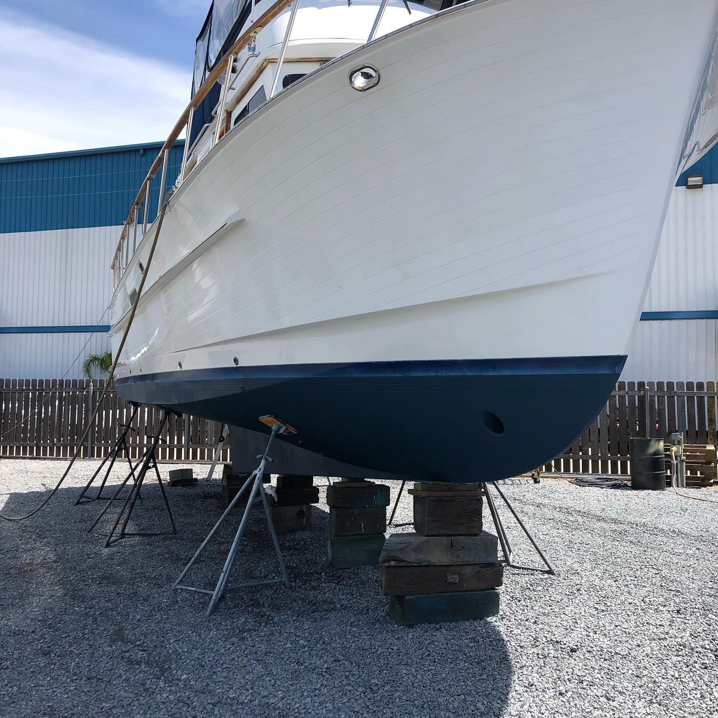 White boat hull on stands in a gravel yard by a blue industrial building