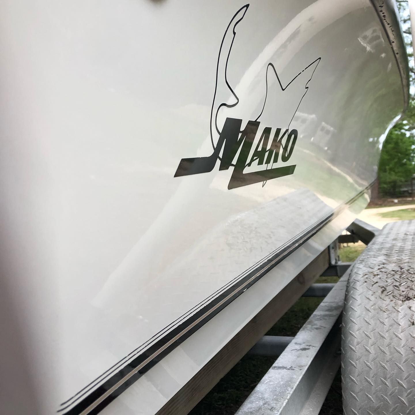 White Mako trailer side with logo, seen close-up beside a diamond-plate step and green trees behind.