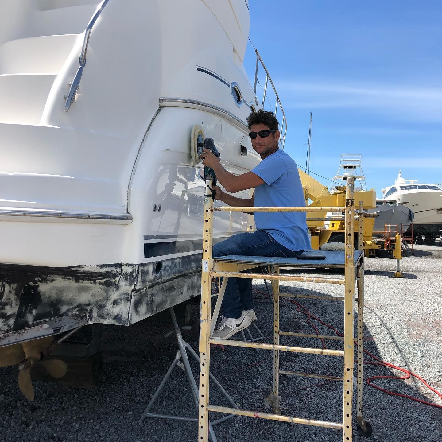 Person sanding a white boat hull while seated on a scaffold in a sunny boatyard