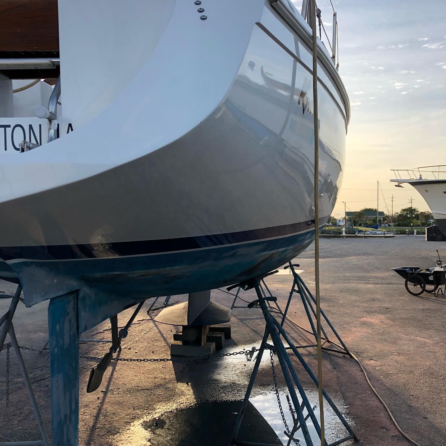 Large white sailboat hull on supports in a marina at sunset, with wet pavement reflecting light.