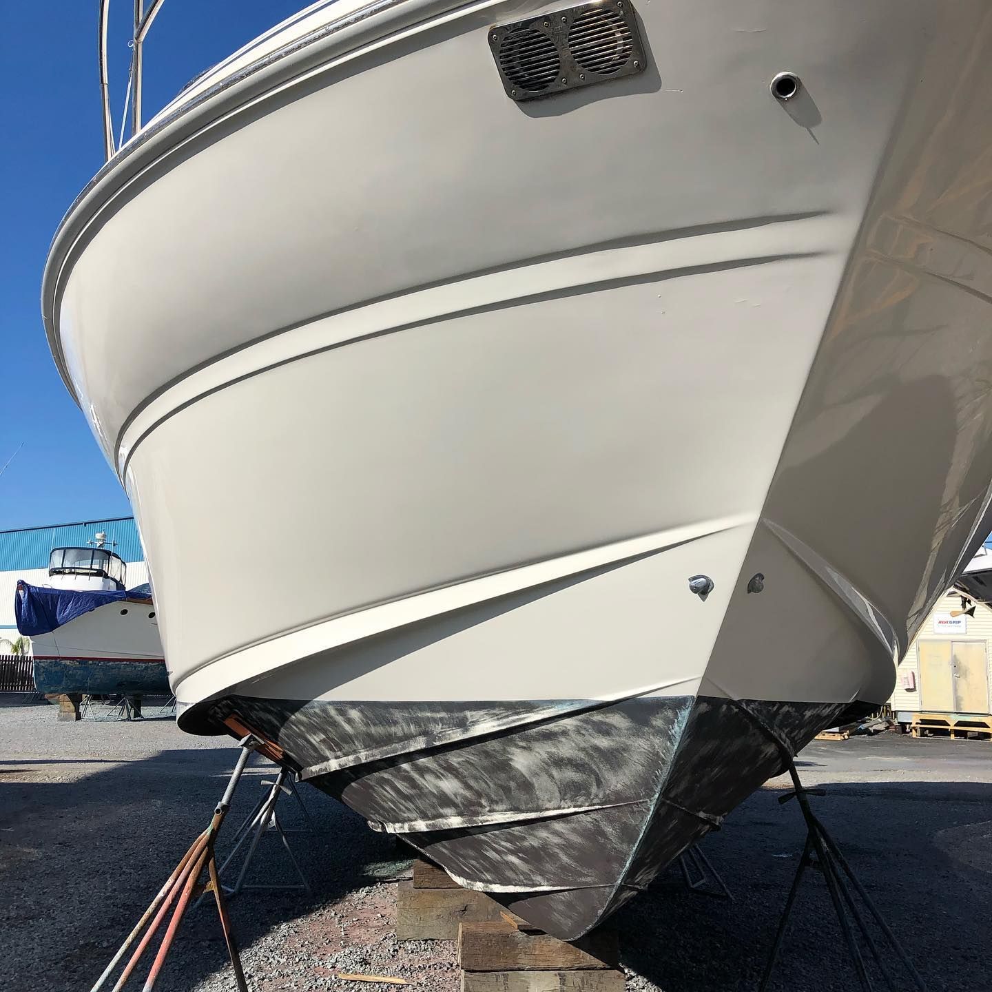 White boat hull with black bottom, propped on stands in a boatyard under a blue sky