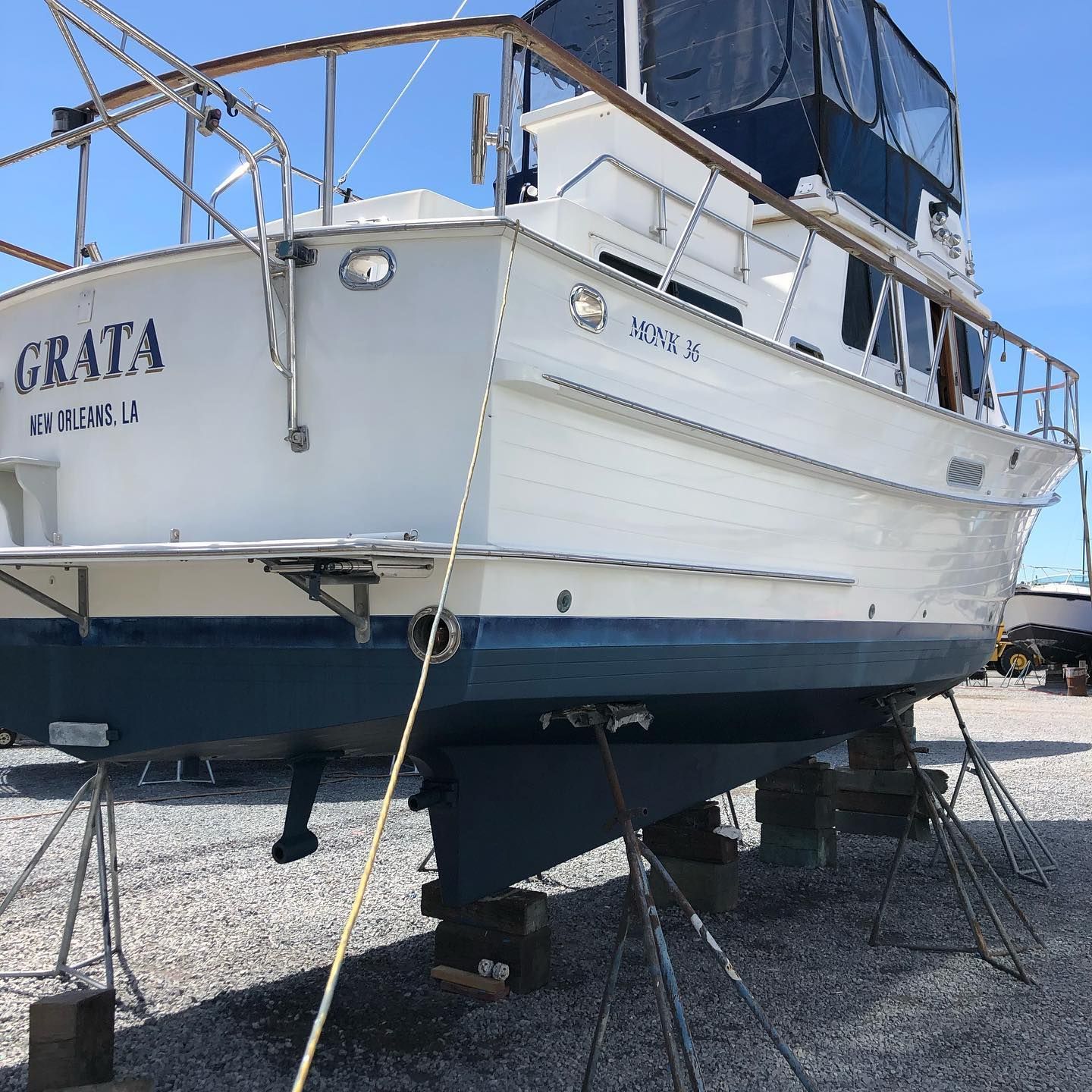 White-and-blue boat on land, viewed from the stern on stands in a gravel yard under a clear blue sky.