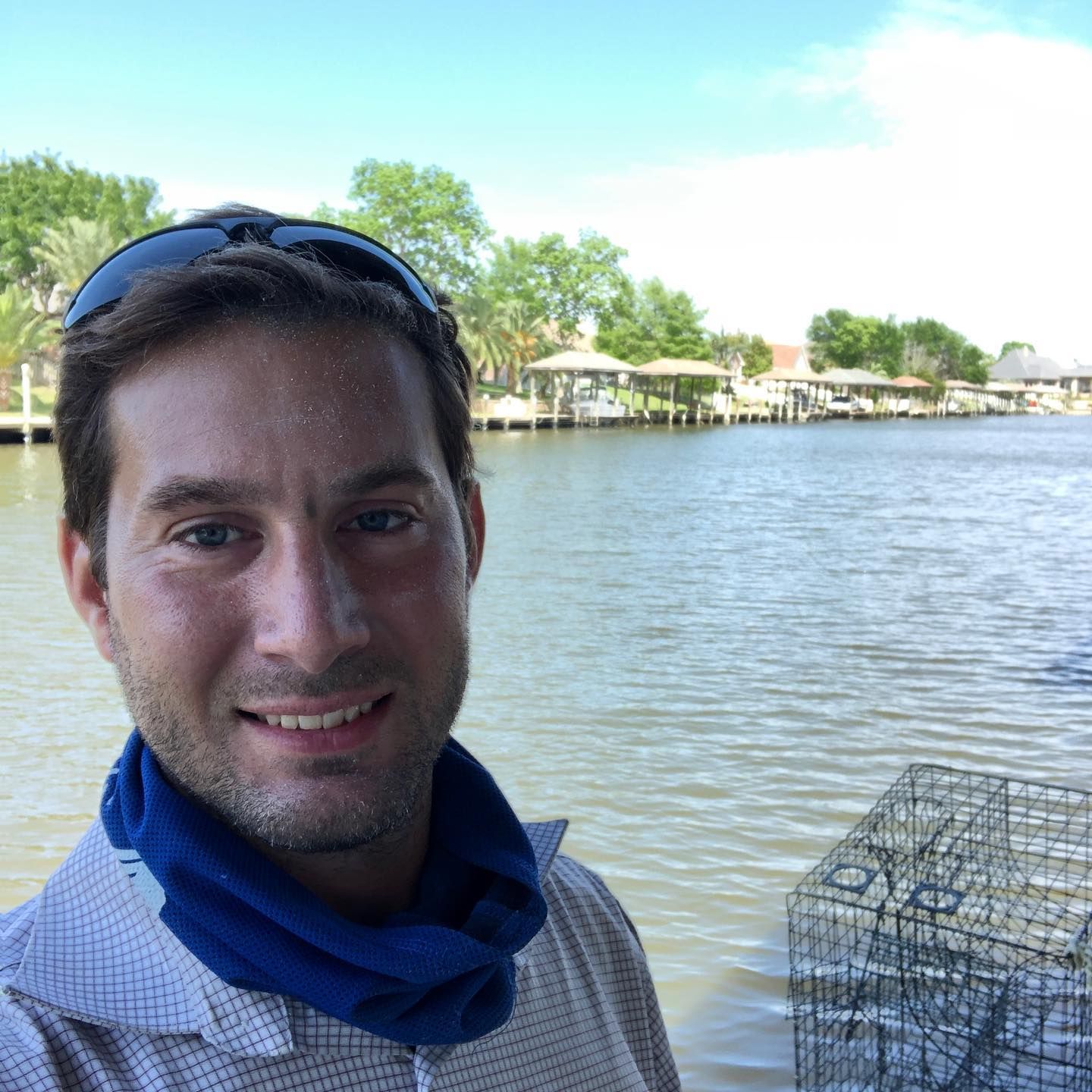 Smiling person in a blue scarf by a lakeside, with trees and a wire crab trap in the foreground.