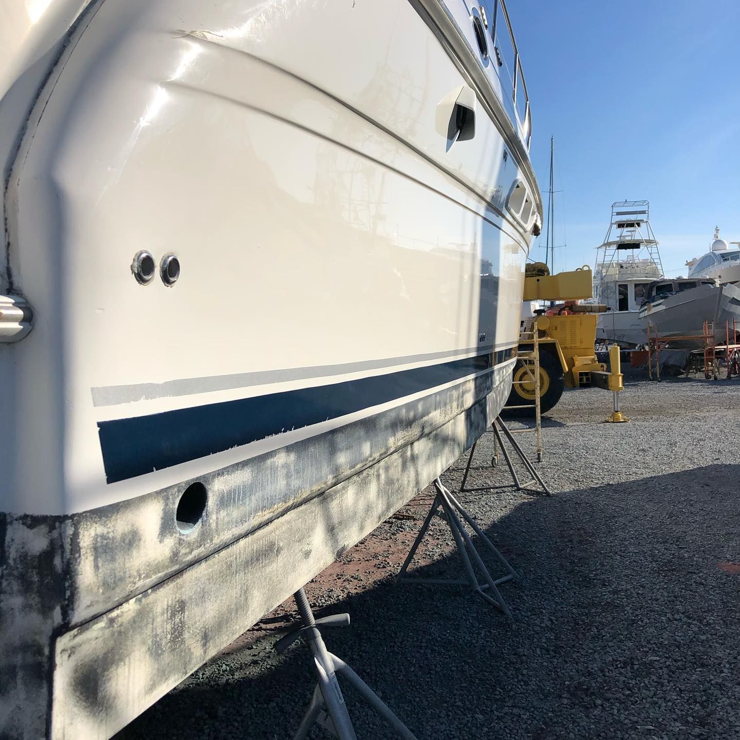 White boat hull on land with blue stripe, viewed from the side in a gravel boatyard.