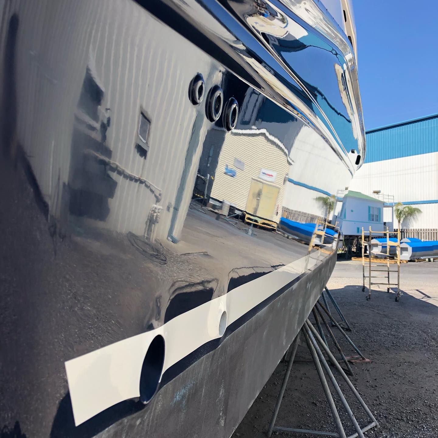 Close-up of a glossy dark-blue boat hull reflecting industrial buildings and sky at a shipyard.