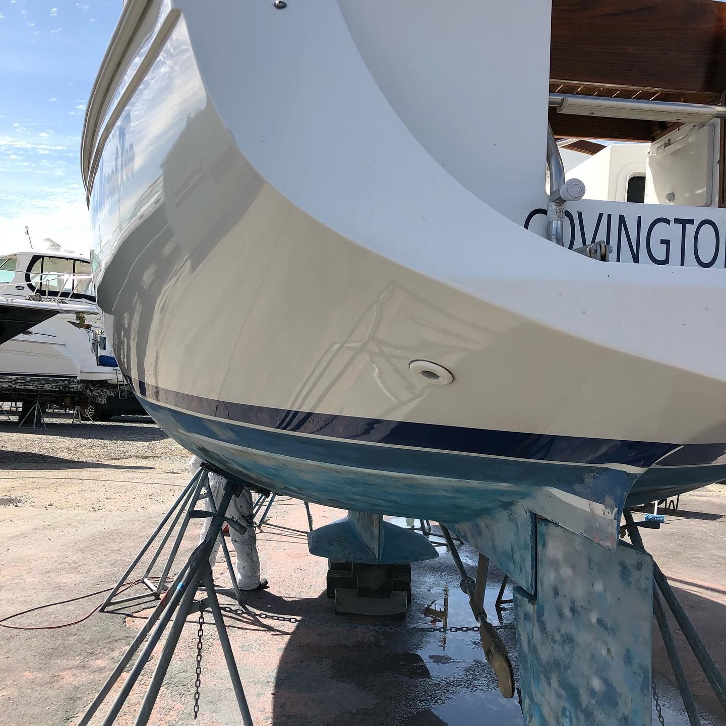 Large white boat hull on metal stands in a shipyard, with blue lower trim and wet ground below.