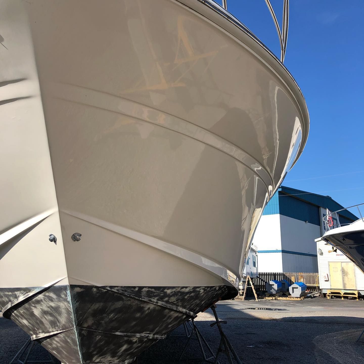 White boat hull in a shipyard under a clear blue sky, with black bottom paint and nearby industrial structures