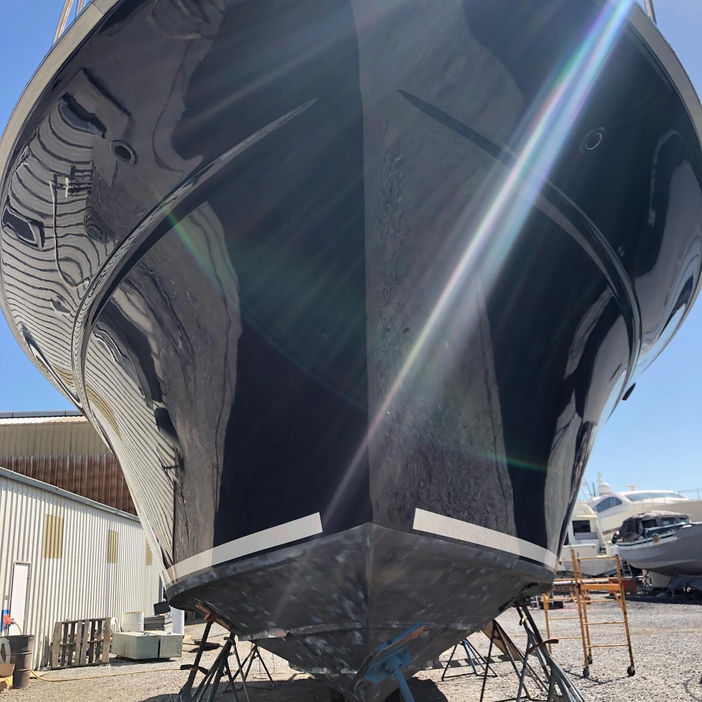 Large black boat hull propped on stands in a boatyard, with bright sun glare and adjacent work trailers