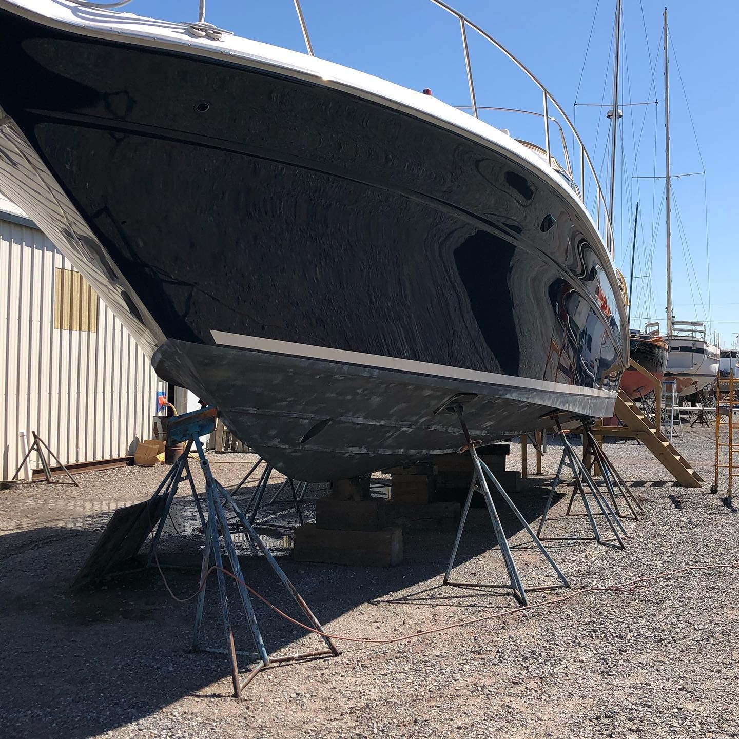 Large sailboat on stands in a boatyard, with a dark hull beside a white shed and gravel ground.