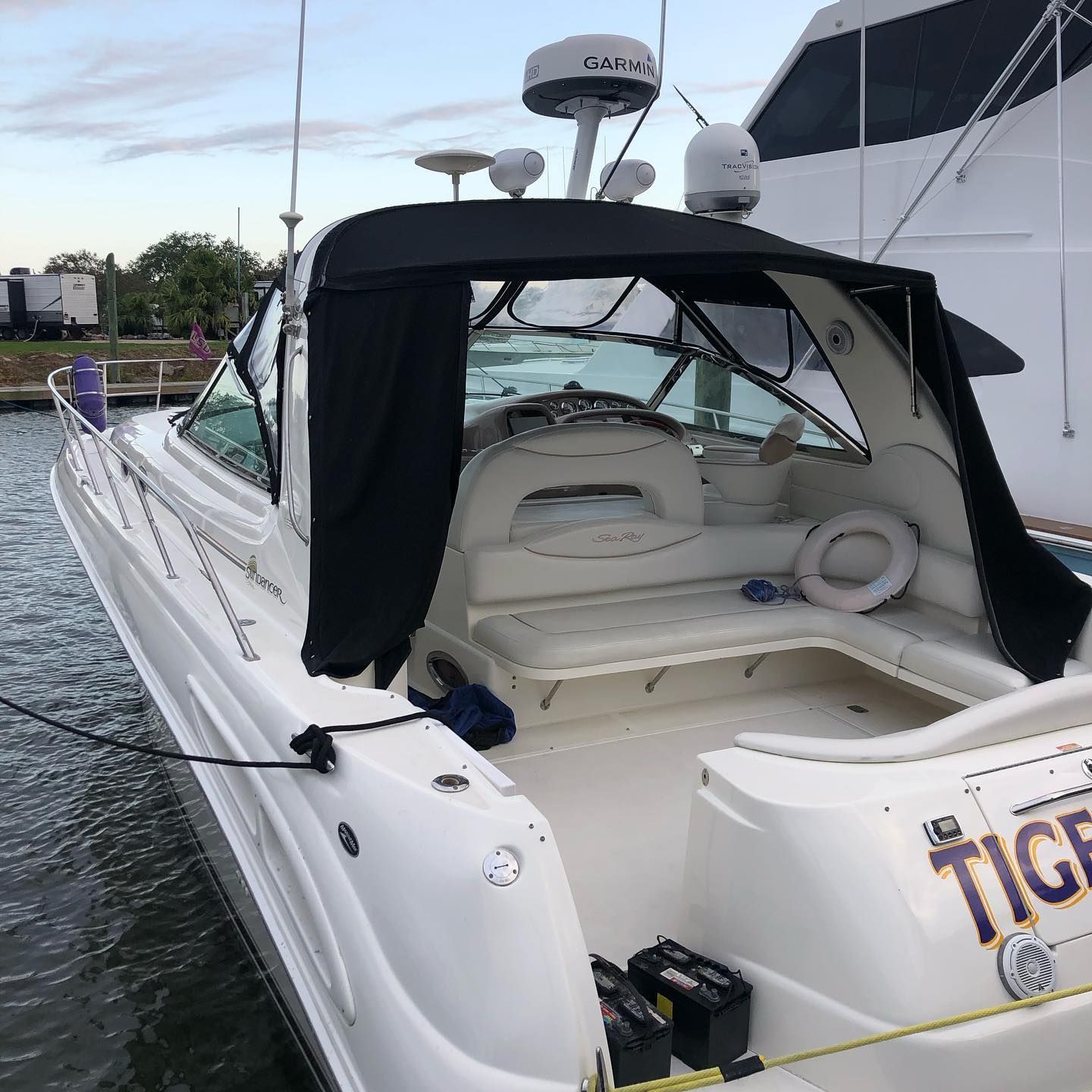 White cabin cruiser with black canopy docked on the water near a larger yacht at dusk