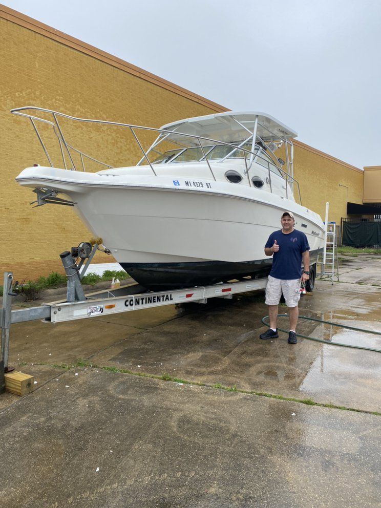 White boat on trailer beside yellow wall, with a man standing in front on a wet concrete lot.