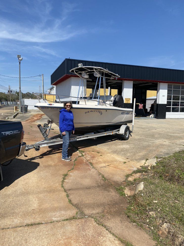 White boat on a trailer with a person beside it outside a garage on a sunny day