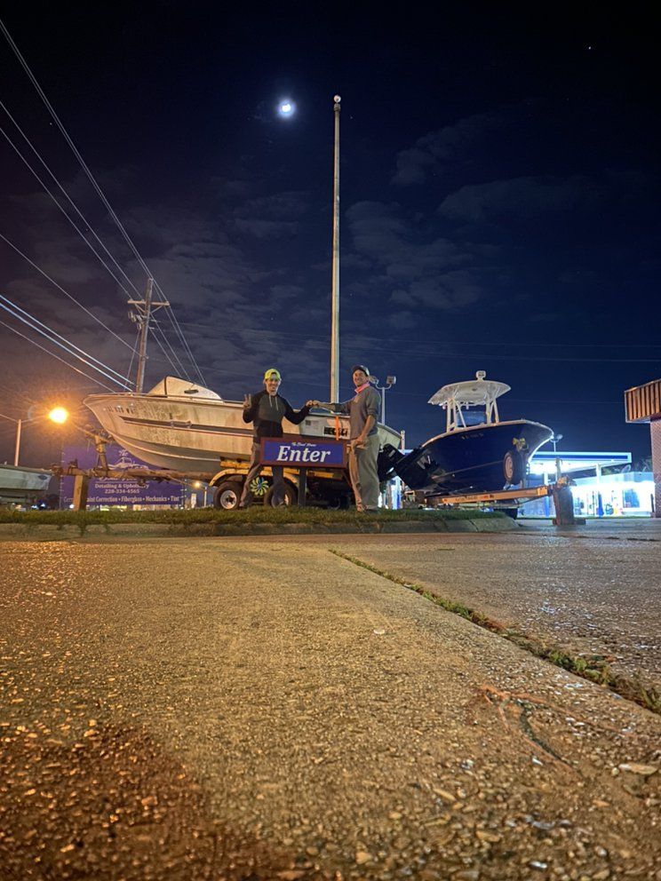 Two people by boats at night near a roadside sign under a moonlit sky.