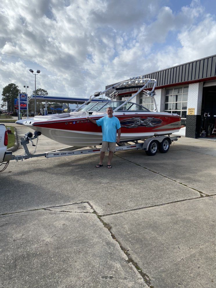 Man in blue shirt stands beside a red-and-white boat on a trailer in a parking lot.
