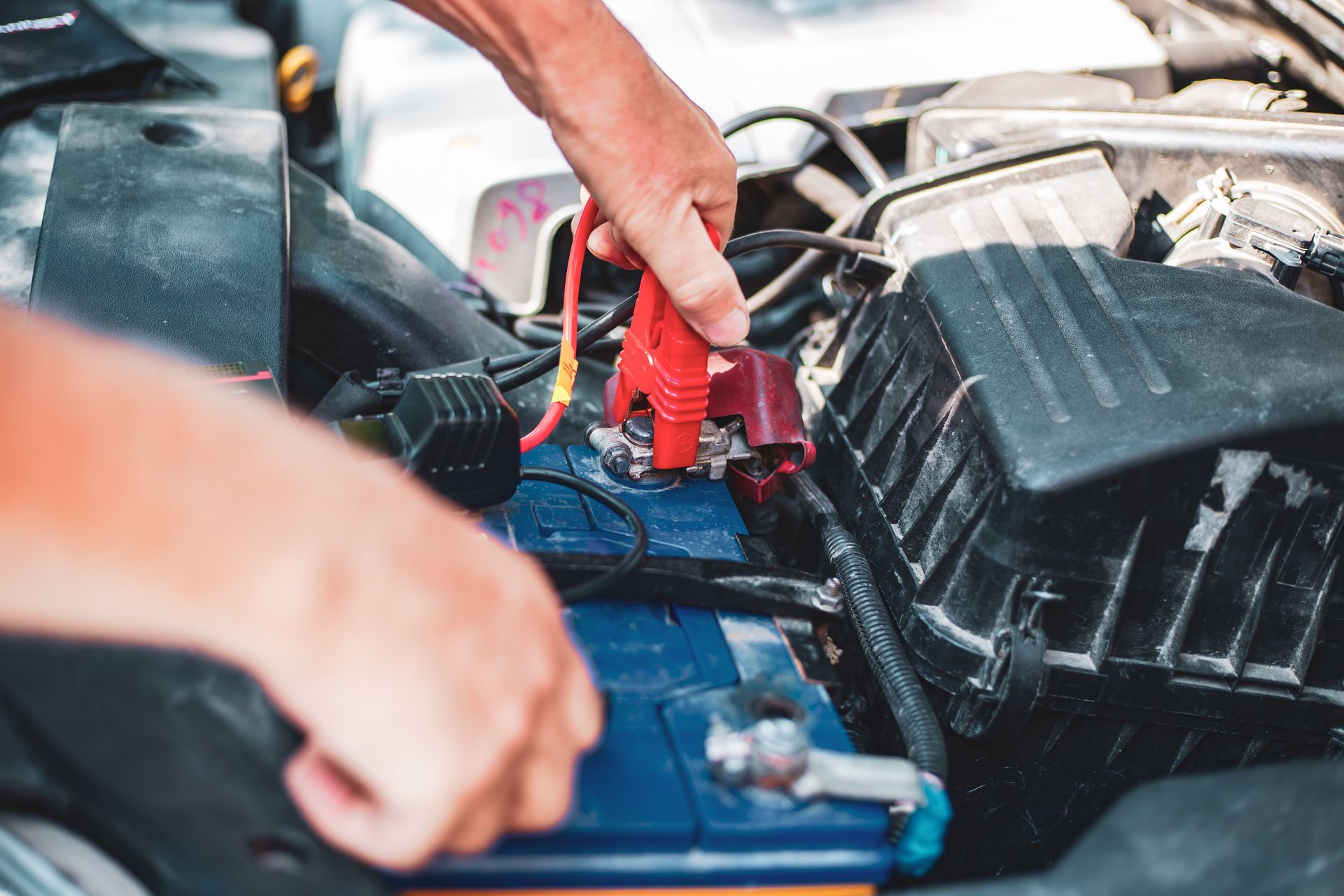 Hands connecting a red jumper cable to a car battery's positive terminal in an engine bay. Hands connecting a red jumper cable to a car battery's positive terminal in an engine bay.