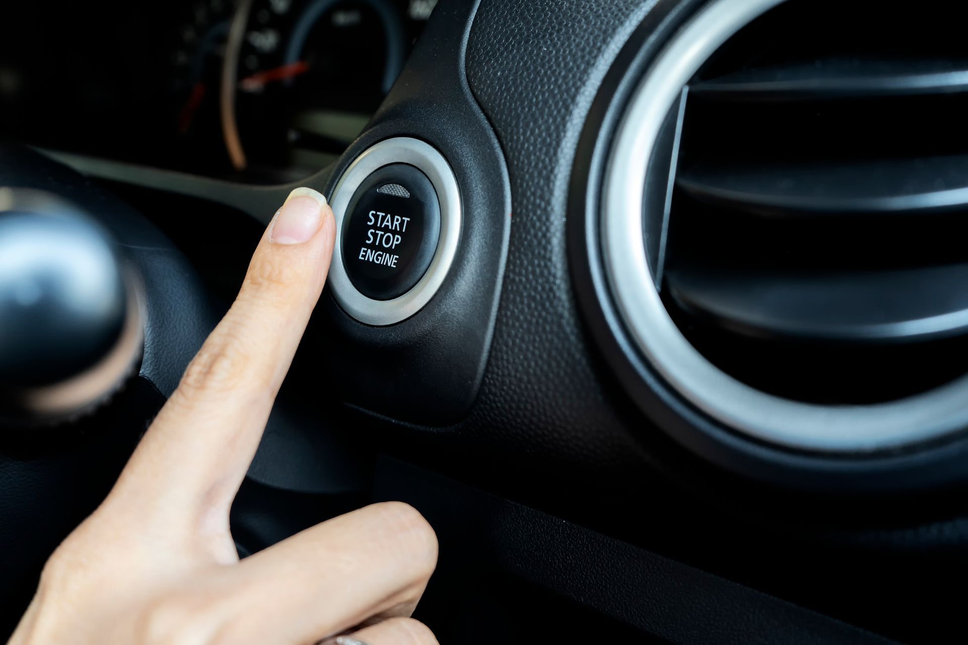 Finger pressing a black car's start/stop engine button, next to an air vent. Finger pressing a black car's start/stop engine button, next to an air vent.
