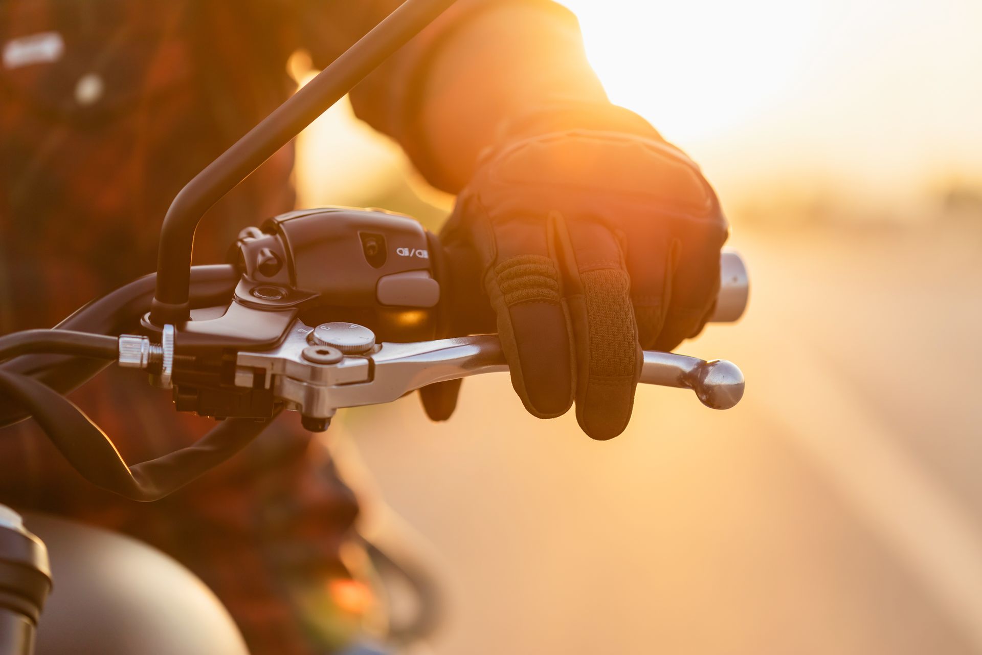 Hand wearing a black glove grips a motorcycle handlebar; sunlight bathes the scene. Hand wearing a black glove grips a motorcycle handlebar; sunlight bathes the scene.