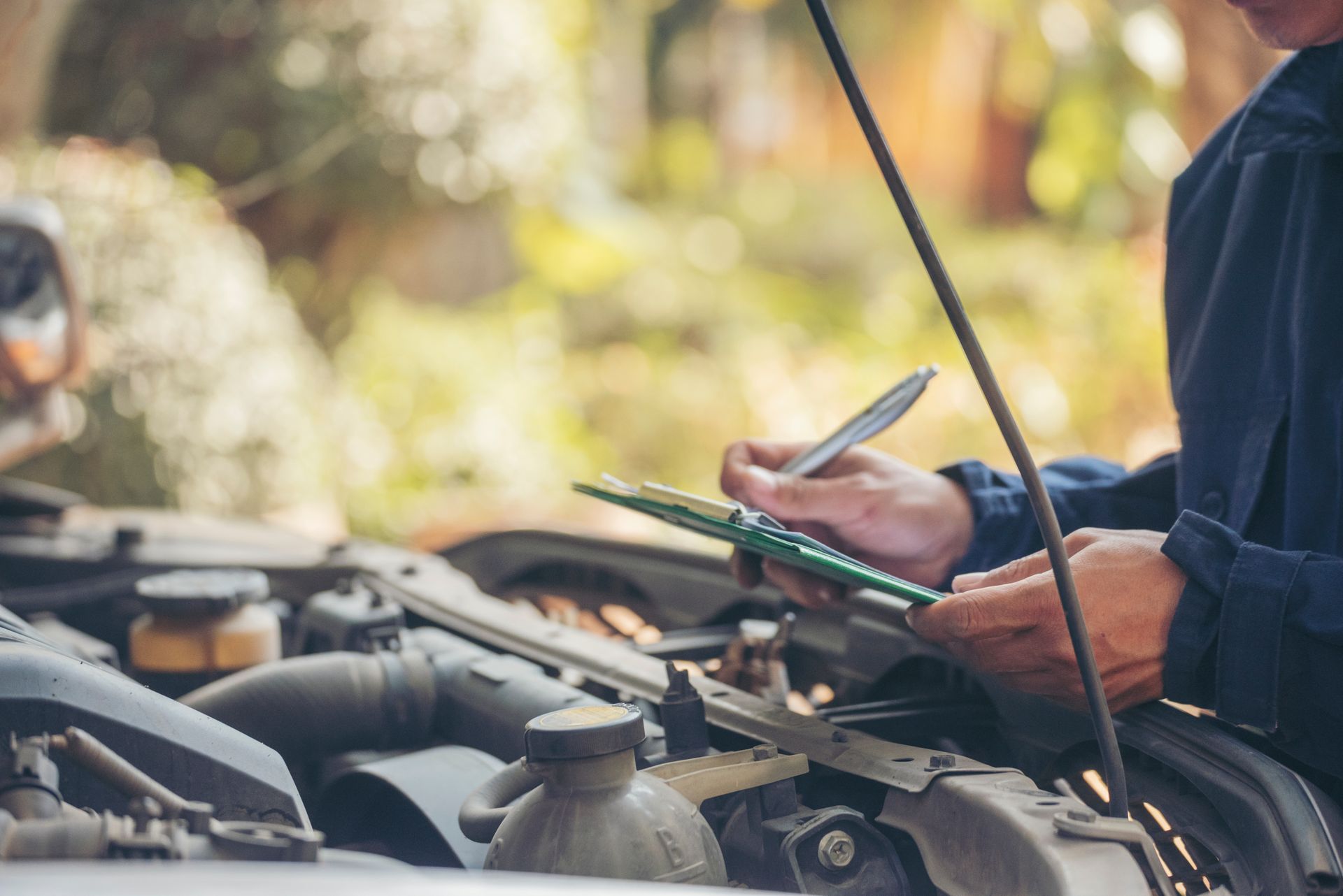 Person inspecting a car engine, taking notes on a clipboard in an outdoor setting.