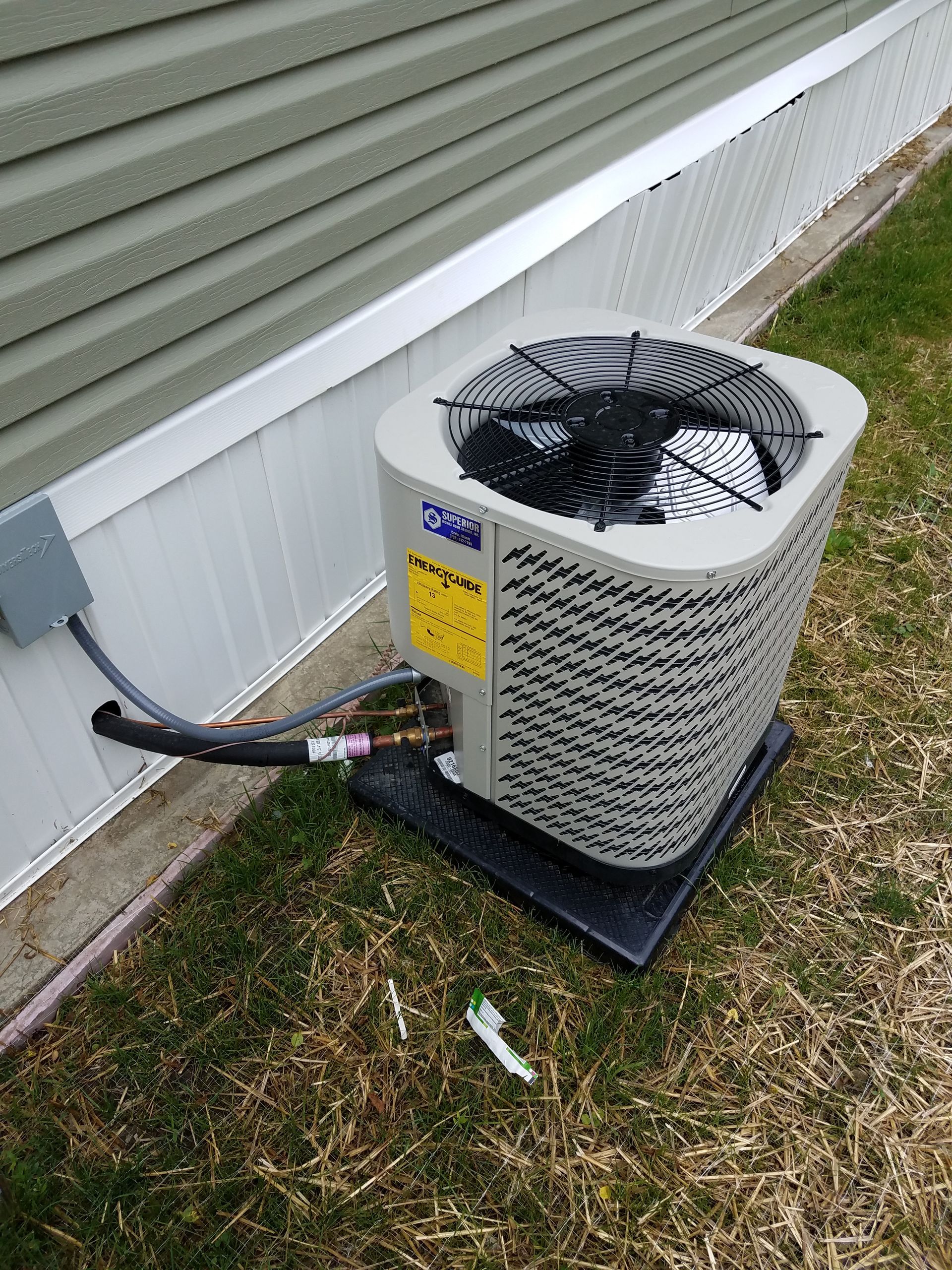 Outdoor air conditioner unit beside a house, with a hose attached and a small tool on the grass.
